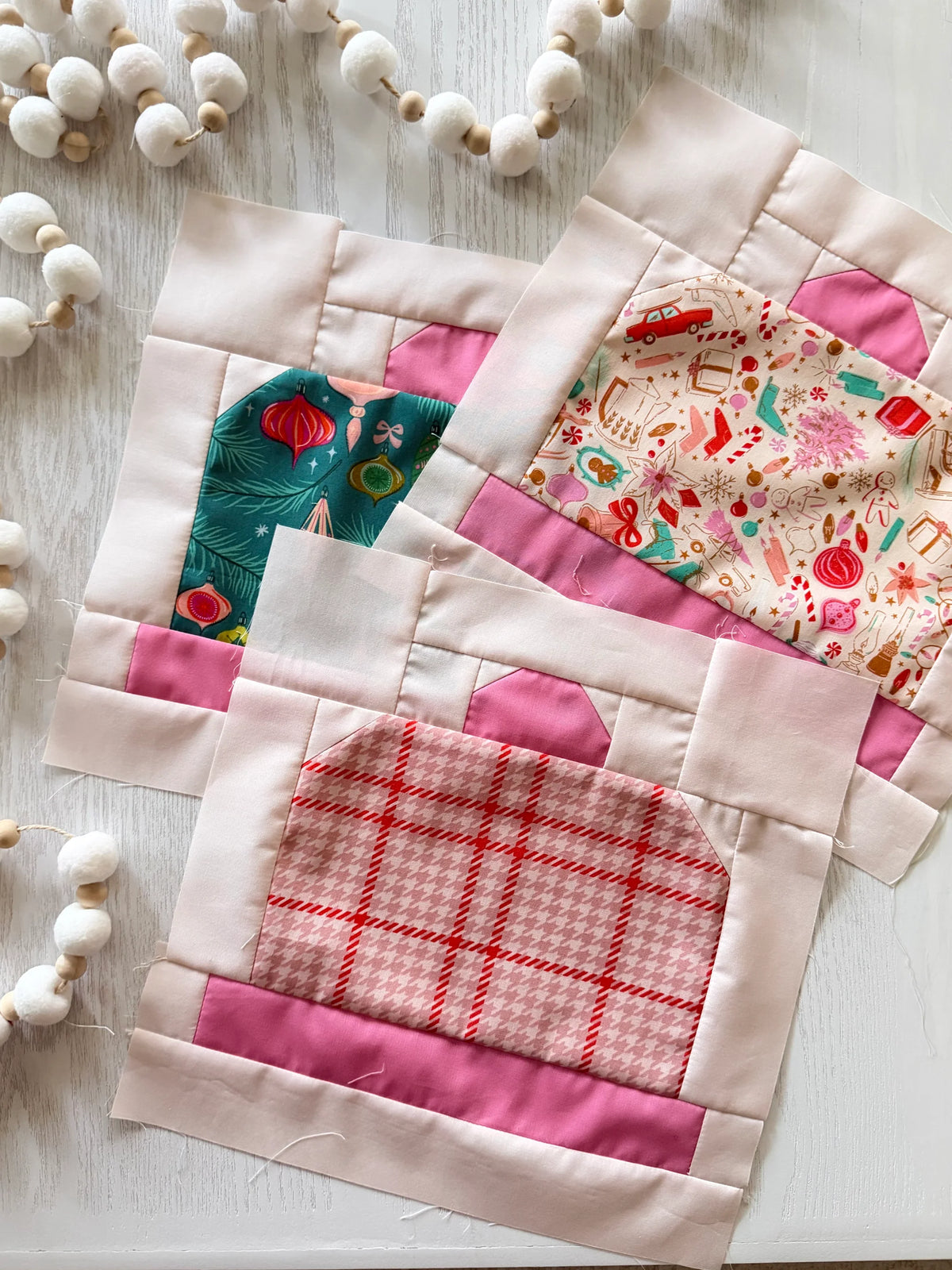 Three handmade quilted fabric blocks with pink borders and colorful patterned centers, arranged on a white wooden surface with decorative white bead garlands.