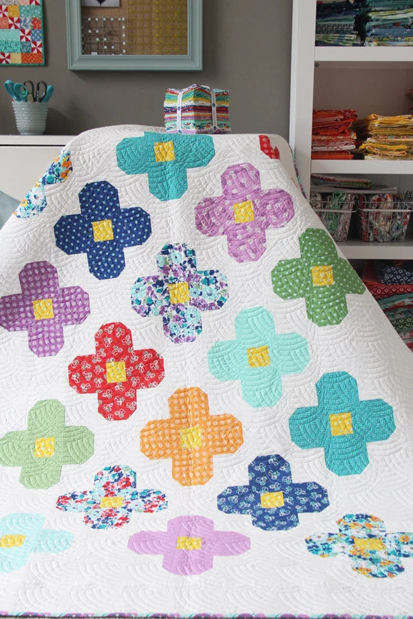 A colorful quilted blanket or throw draped over a chair in a craft room, featuring large patchwork flower shapes in teal, purple, red, orange, green, blue, and pink on a white background.