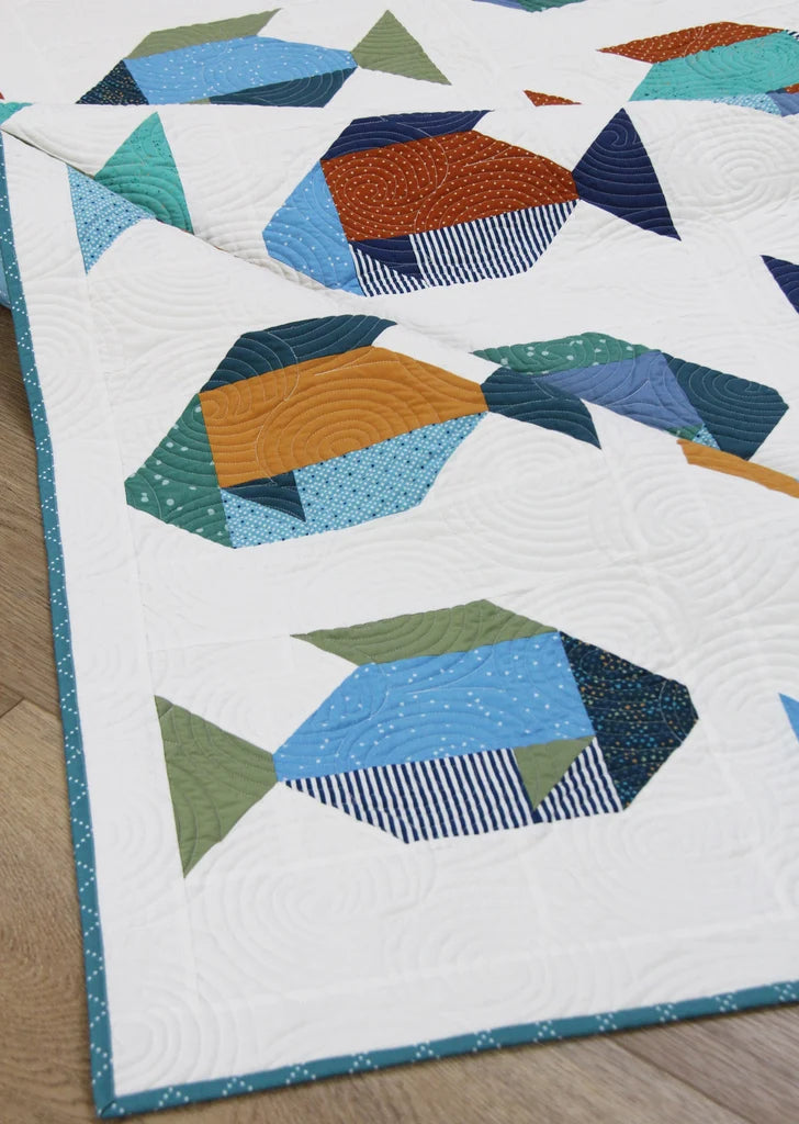 A close-up of a quilted textile featuring geometric fish motifs in blue, teal, orange, green, and navy on a white background, laid on a light wood floor.
