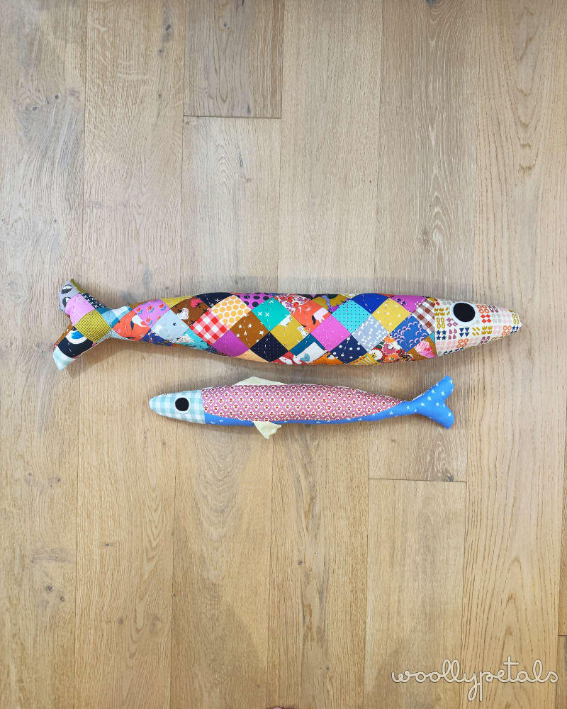 Two colorful fish-shaped toys on a wooden floor with 'woollypotts' branding.