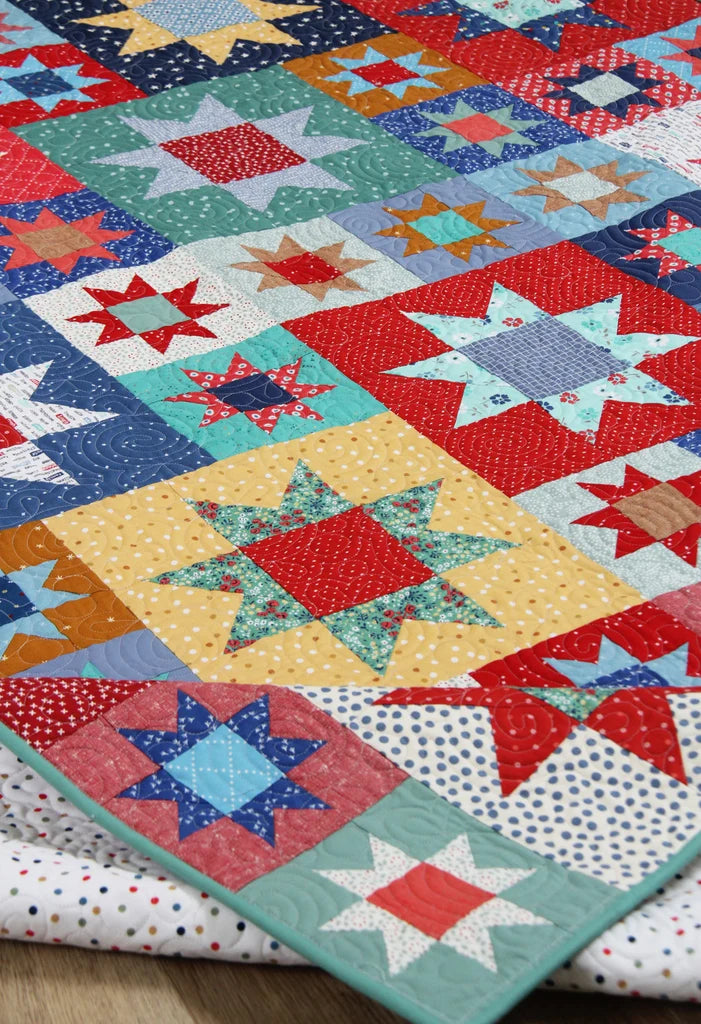 A close-up of a colorful patchwork quilt with star and geometric block patterns in red, blue, teal, yellow, and white fabrics, with folded patterned quilts visible underneath on a wooden surface.