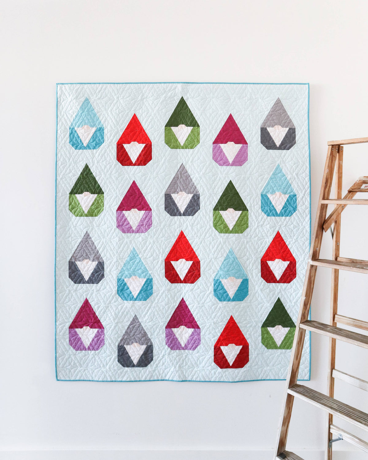 A quilted wall hanging or blanket featuring a grid of colorful gnomes inside teardrop shapes, displayed on a white wall next to a wooden ladder.