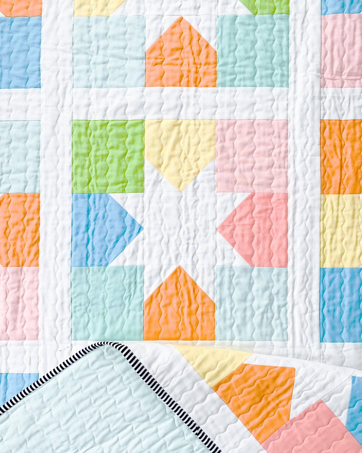 A close-up of a quilted fabric with a pastel geometric pattern of triangles and squares in light blue, mint, pink, yellow, green, and orange on a white background.