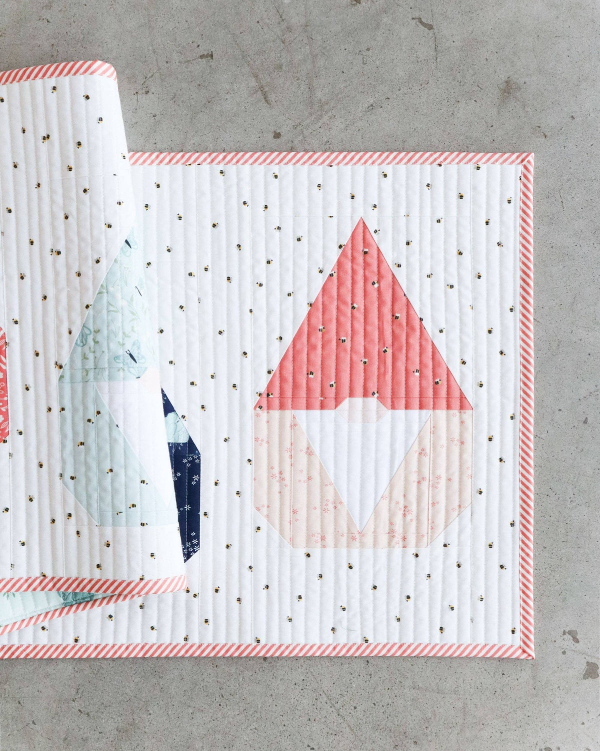 A quilted fabric panel or quilt with a gnomehouse motif, shown partially rolled up on a gray concrete surface. The fabric features a white base with tiny black dots, a coral-pink gnomehouse, and other geometric shapes in light blue and navy.