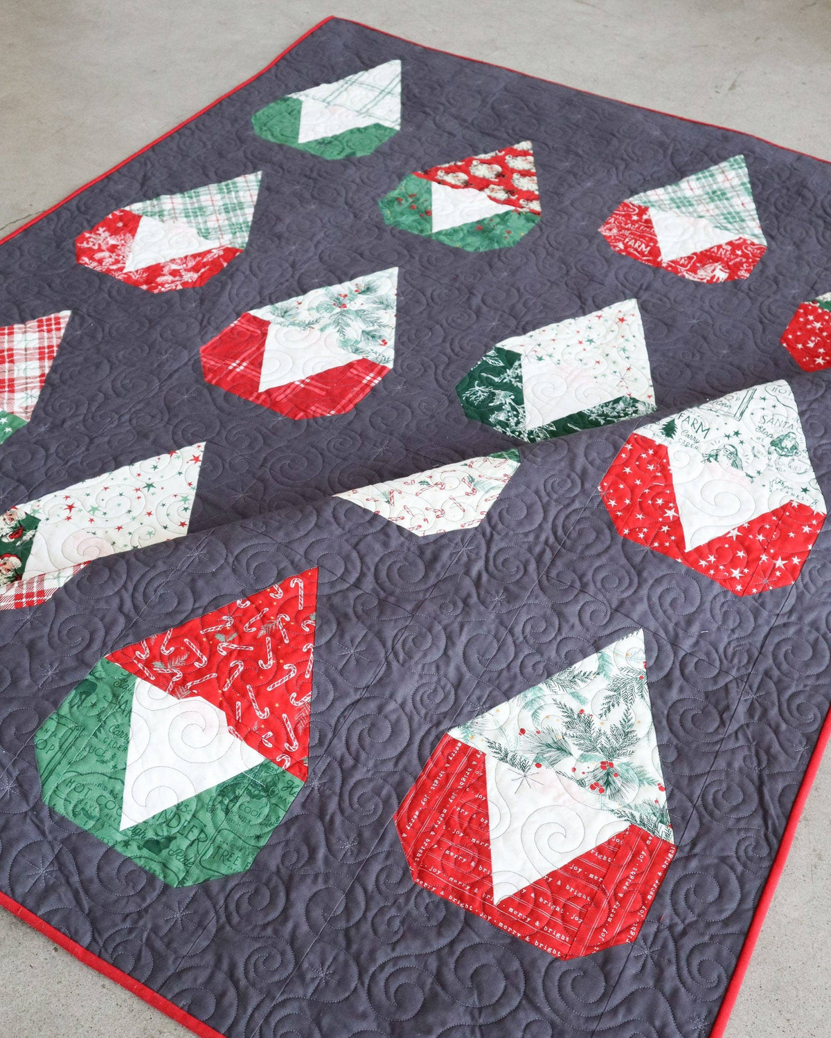 A handmade quilted blanket or throw featuring a dark gray background with a pattern of red, green, and white geometric holiday shapes, laid out on a neutral concrete floor.