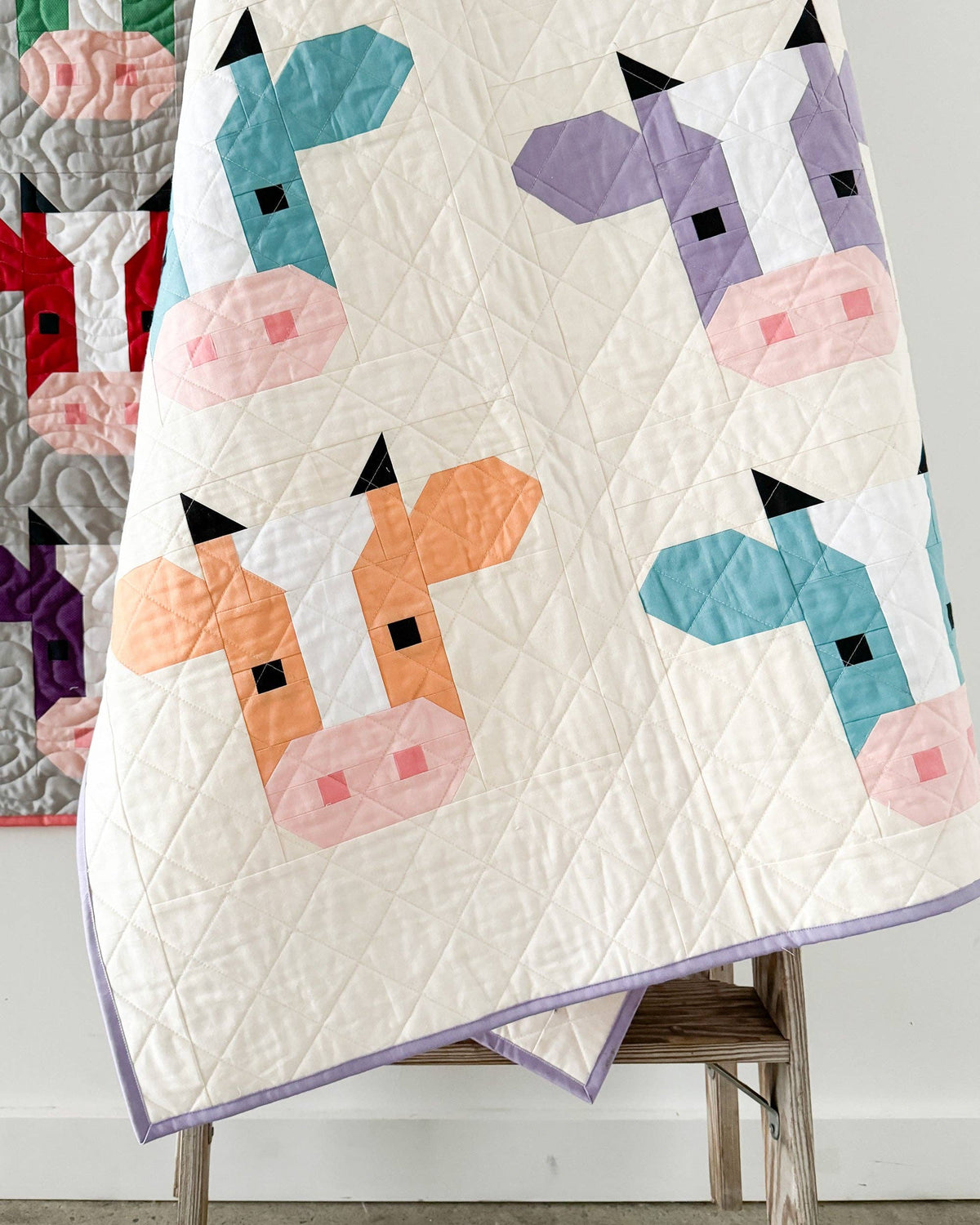 A quilted baby blanket featuring a playful patchwork design of stylized cow faces in teal, purple, orange, and pink on an off-white background, draped over a wooden chair.