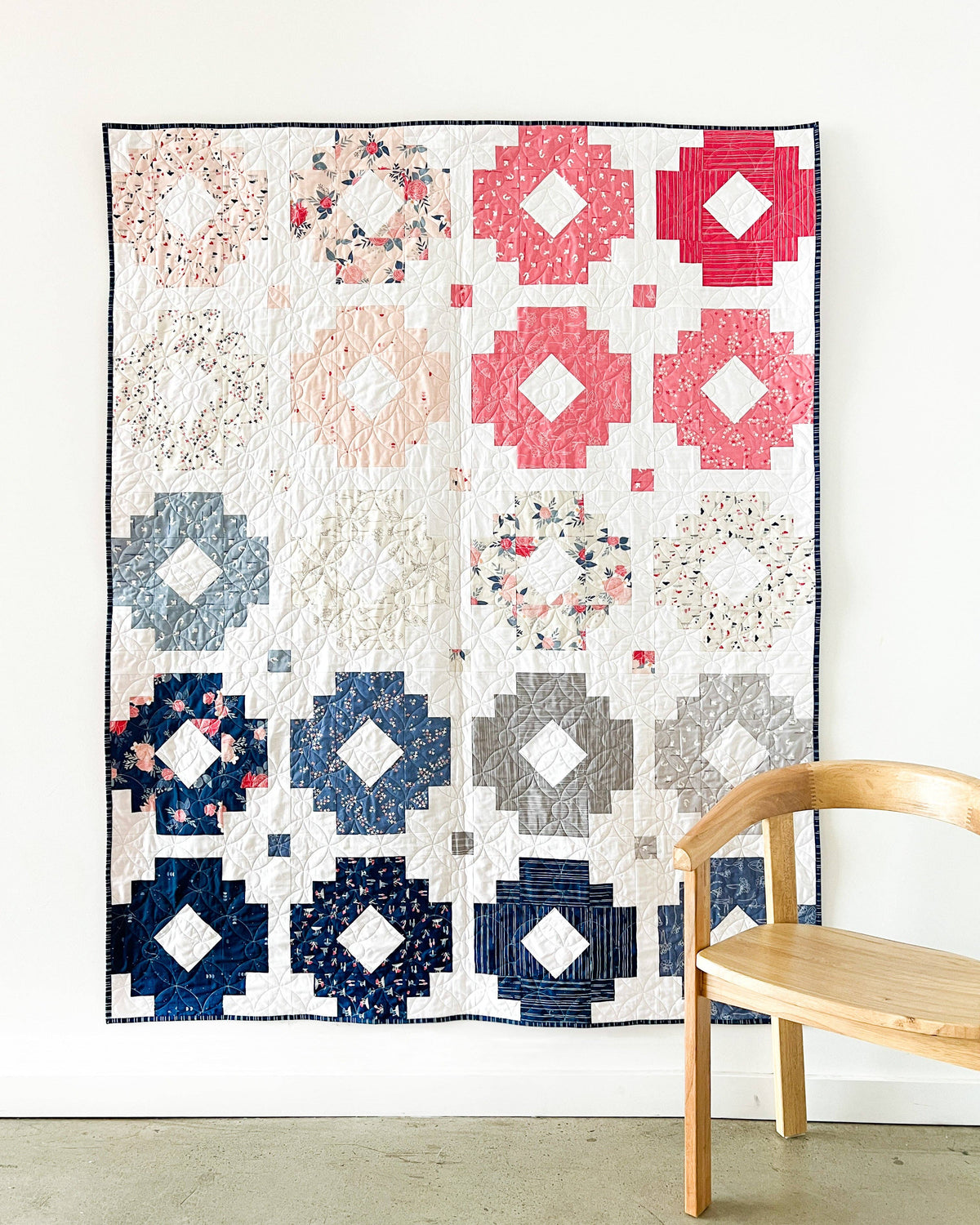 A handmade patchwork quilt hanging on a white wall, featuring a grid of geometric and floral circular blocks in navy, pink, blue, gray, and cream. A light wood chair is partially visible in the foreground on the right.