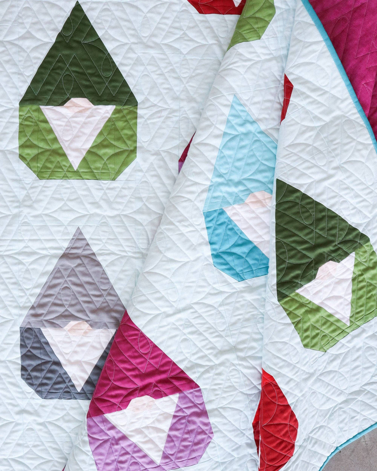 A close-up of a quilted textile with geometric shapes in green, light blue, pink, red, gray, and purple on a white background.