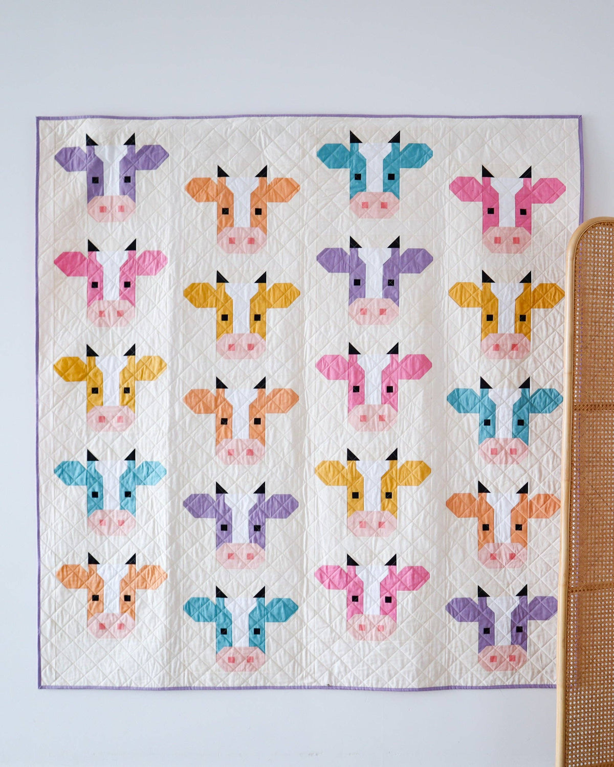 A square quilted textile featuring a repeating pattern of stylized cow faces in multiple pastel colors, including pink, purple, blue, orange, and yellow, displayed against a light wall with part of a woven chair visible on the right.
