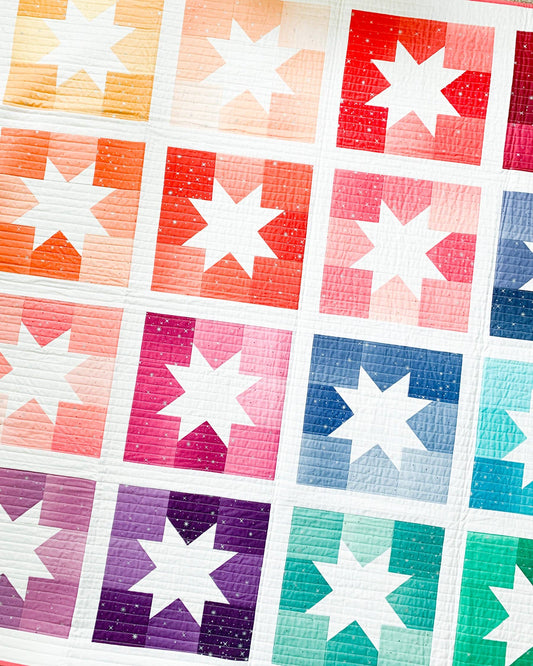 A close-up of a quilted fabric featuring a grid of colorful star blocks in orange, red, pink, blue, purple, and green on a white background.