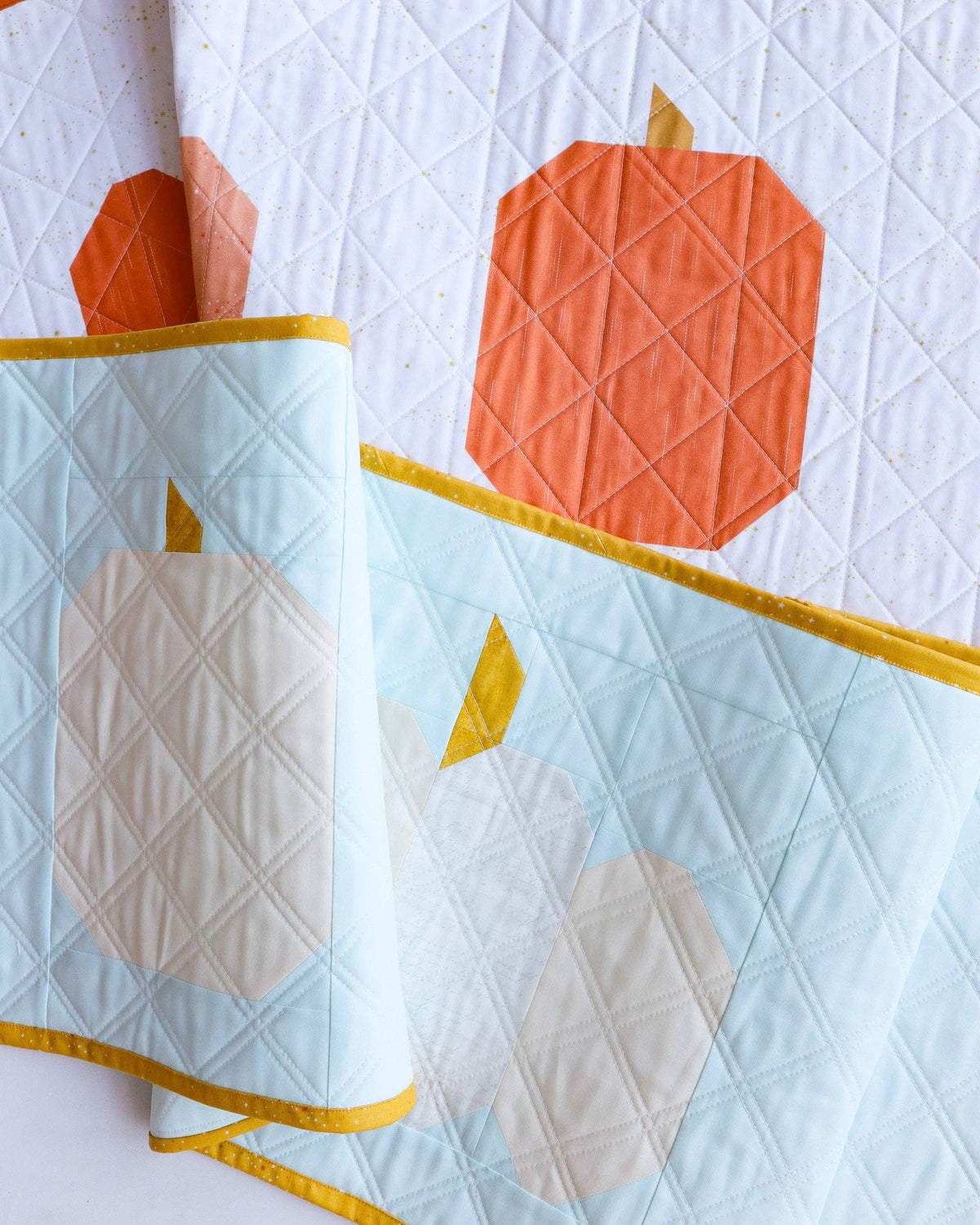 A close-up of a quilted textile featuring large orange and cream pumpkin motifs on a pale blue background, with yellow edging and a white quilted surface behind it.