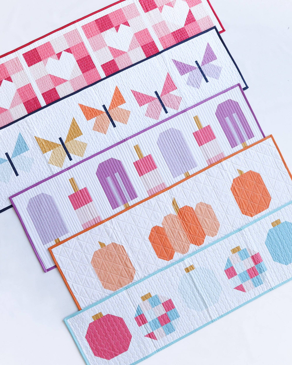 Five colorful quilted fabric table runners arranged diagonally on a white background, featuring geometric patterns of butterflies, popsicles, pumpkins, ornaments, and hearts.
