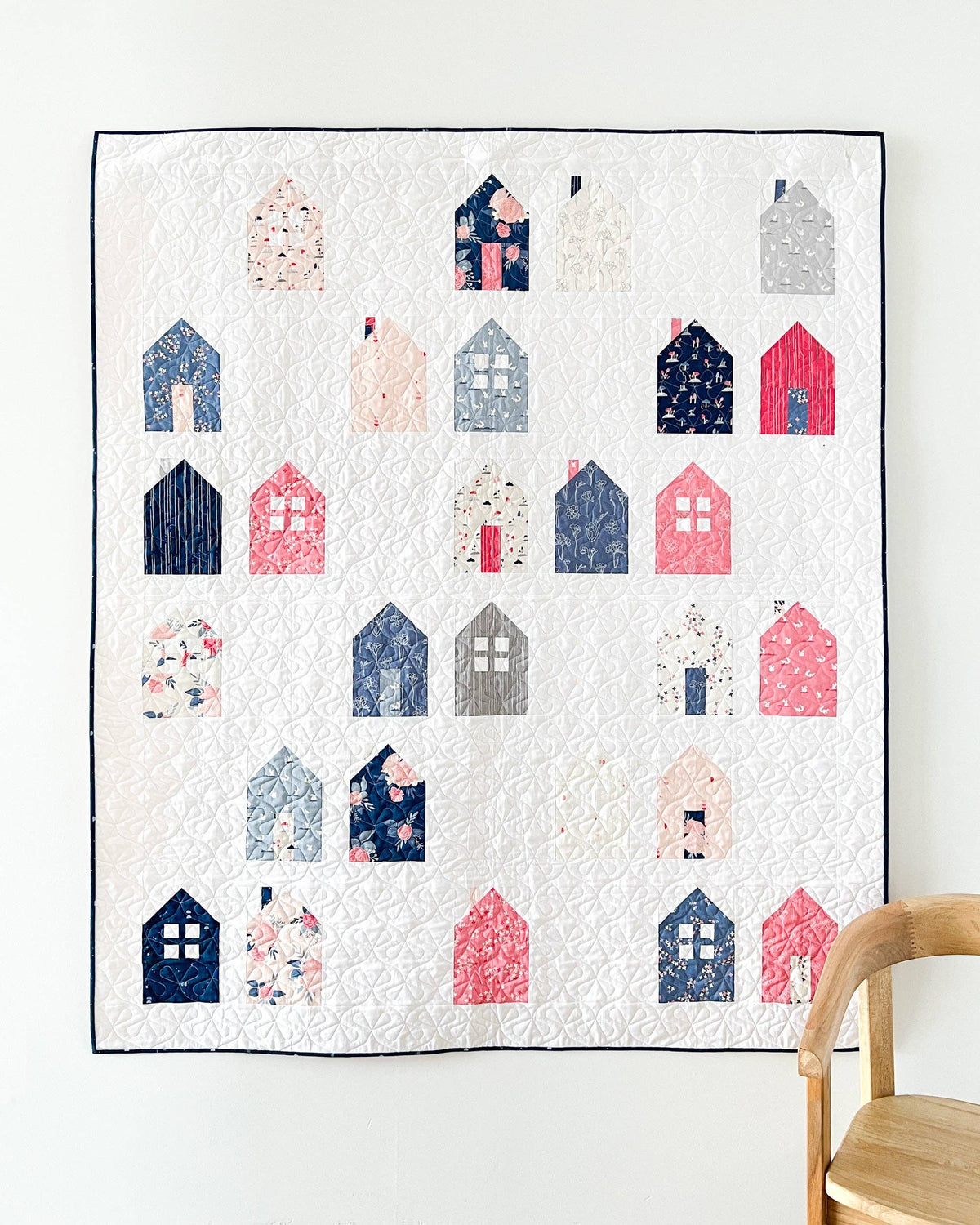 Quilt with house pattern on a white wall with a wooden chair.