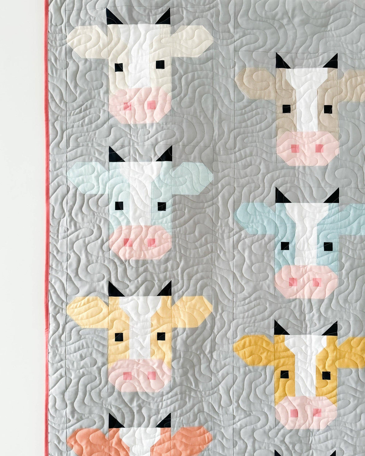 A close-up of a quilted fabric featuring a repeating pattern of stylized cow faces in pastel colors including cream, light blue, yellow, peach, and pink on a light gray background.