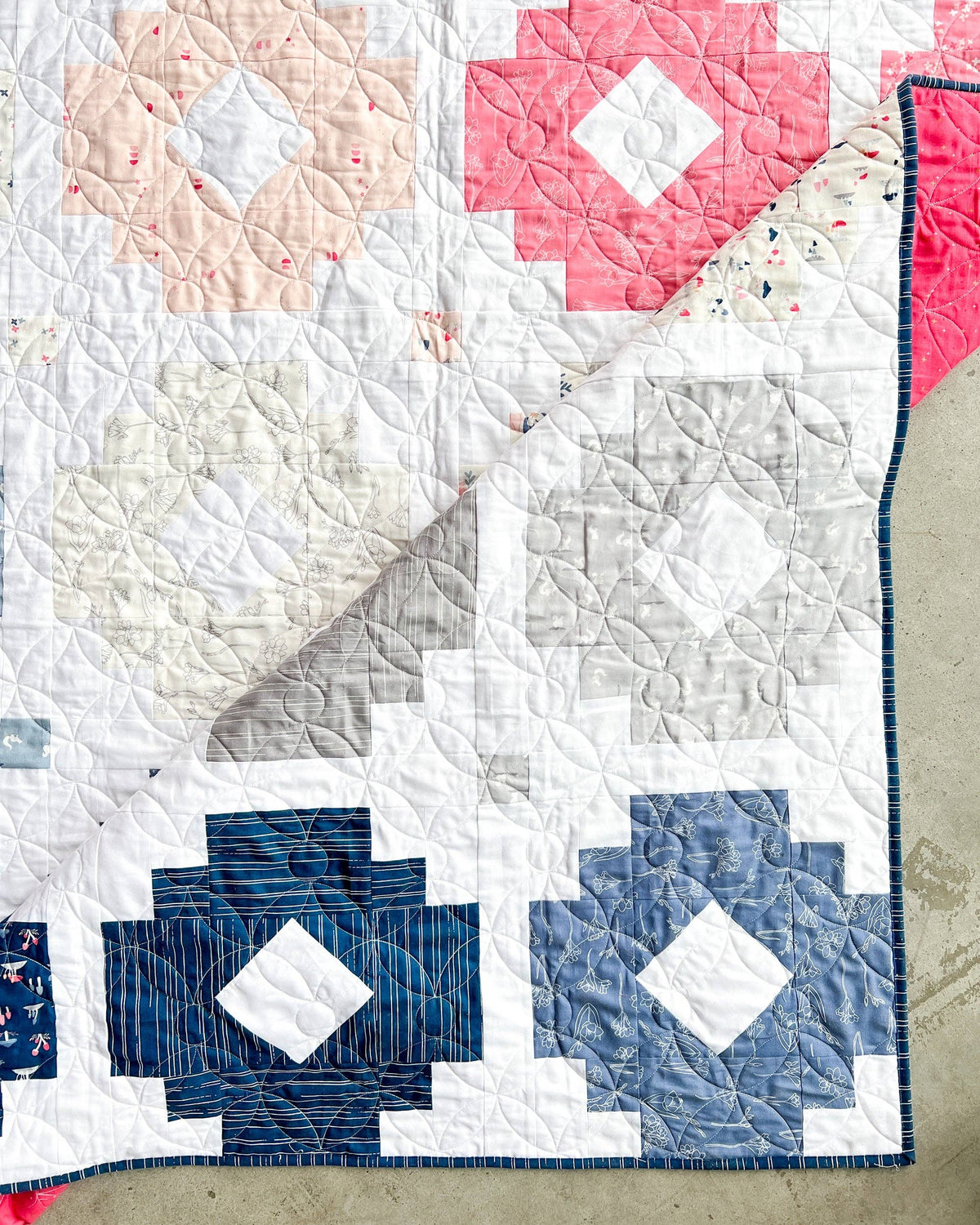 A handmade quilted patchwork blanket featuring geometric block patterns in pink, blue, gray, and white, with a folded corner revealing the underside.