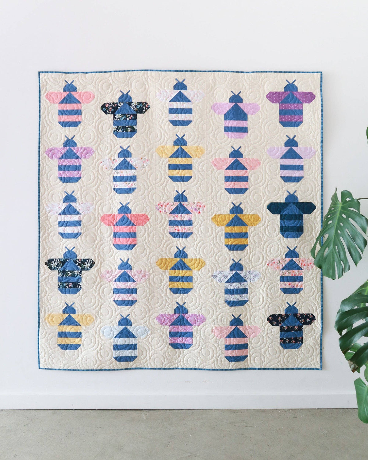 A quilted wall hanging or blanket featuring rows of stylized bumblebee motifs in blue, pink, yellow, and black on a cream background, displayed against a white wall with a green plant leaf at the right edge.