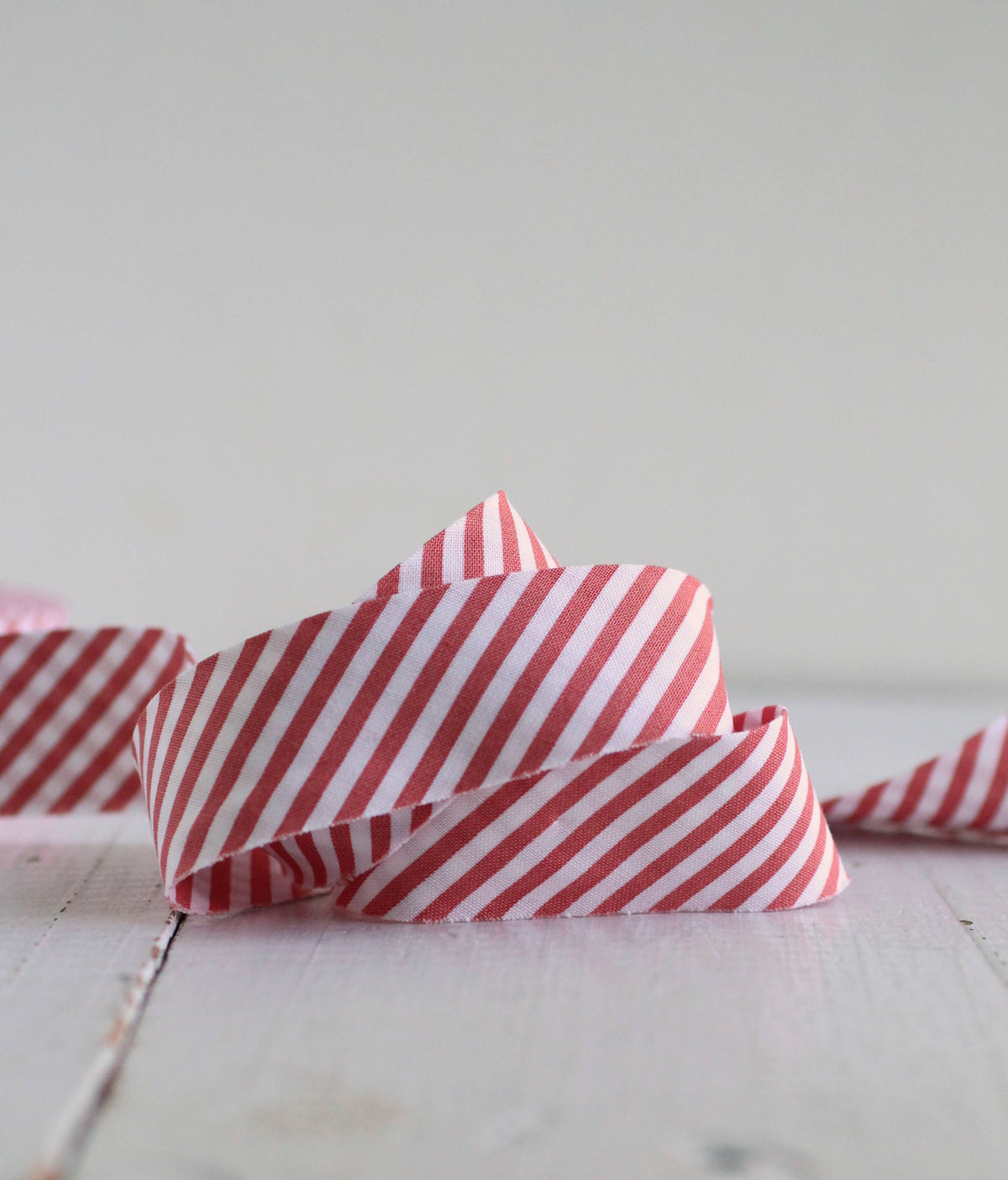 A close-up of a red and white striped bias quilt binding, styled like candy canes, coiled on a light-colored wooden surface against a soft, neutral background.