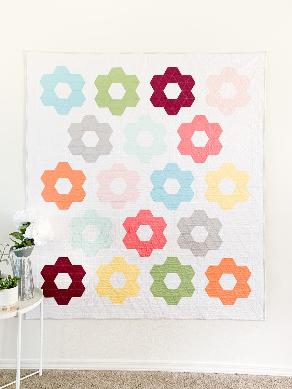 Colorful floral quilt hanging on a white wall with a small table and plant in the foreground.