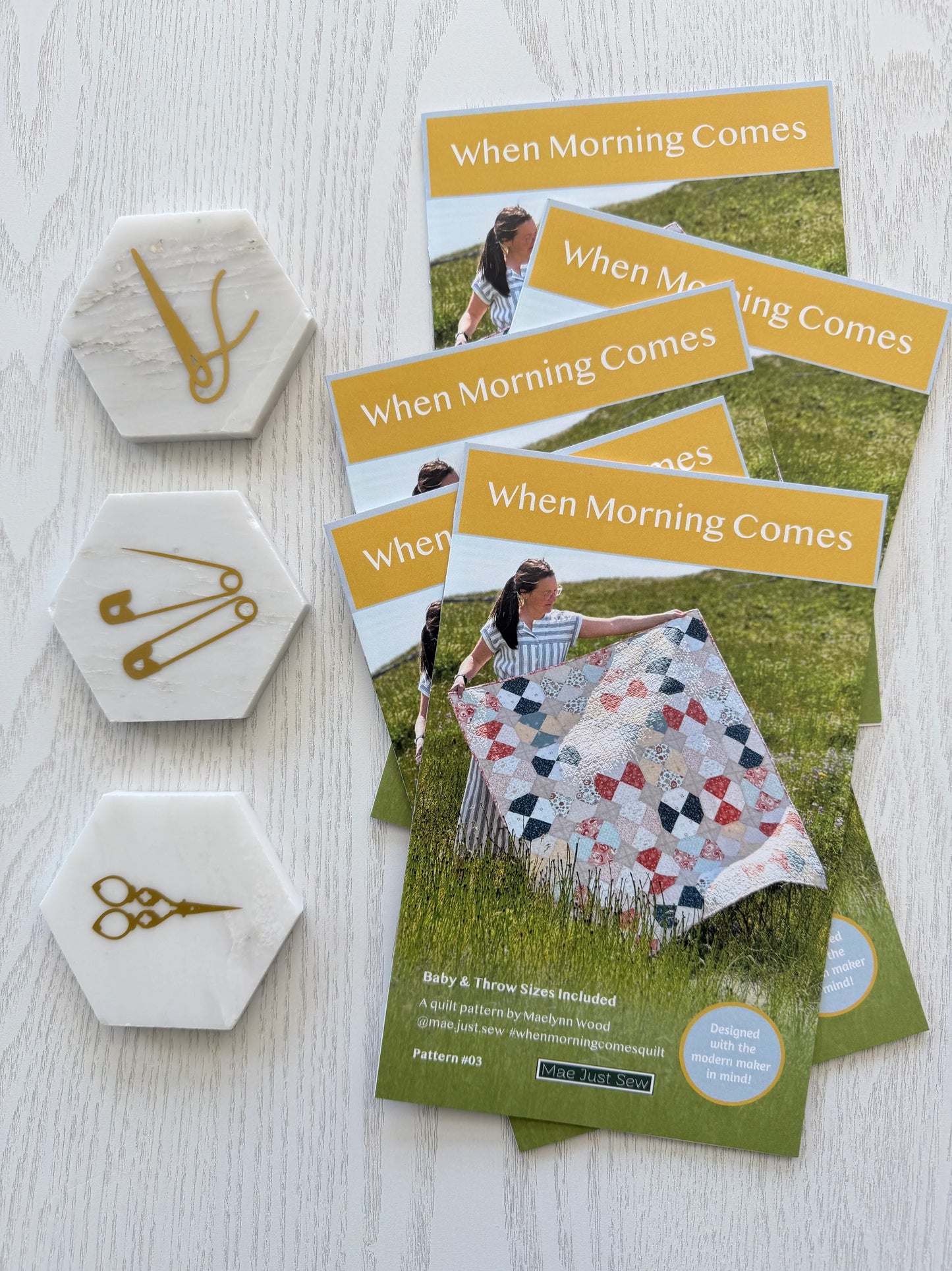 Quilt pattern booklets titled 'When Morning Comes' with hexagonal coasters featuring sewing tools on a light wooden surface.