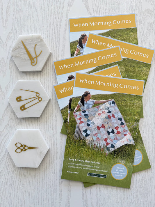 Quilt pattern booklets titled 'When Morning Comes' with hexagonal coasters featuring sewing tools on a light wooden surface.