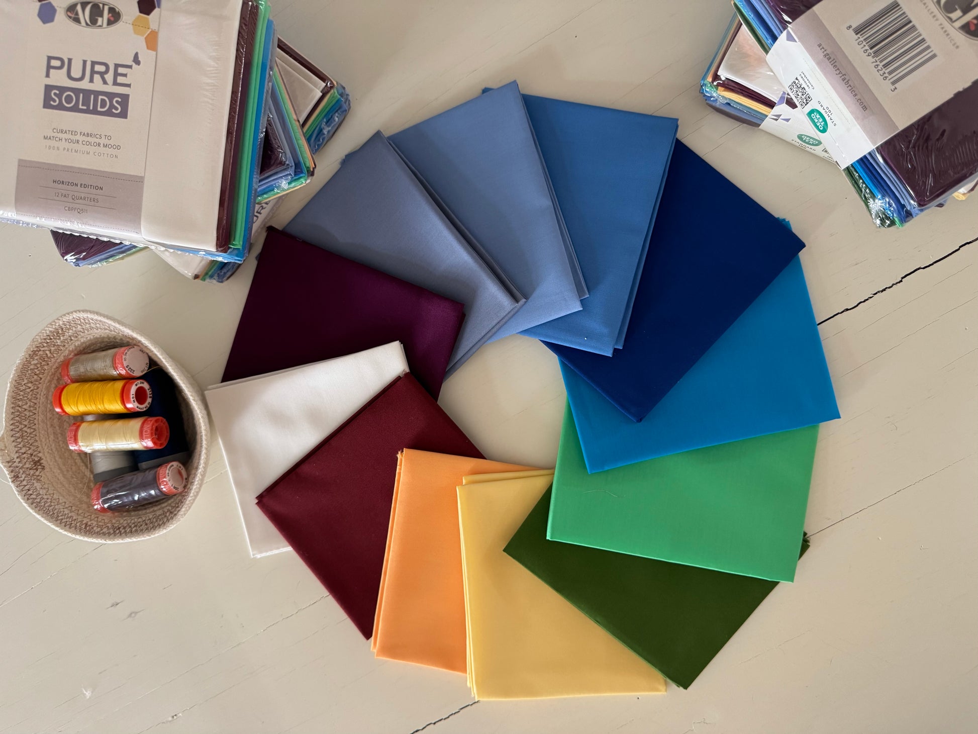 A circular arrangement of folded fabric swatches in multiple colors, including blue, purple, white, burgundy, orange, yellow, green, and teal, displayed on a light wooden surface with sewing thread spools and fabric packages around them.