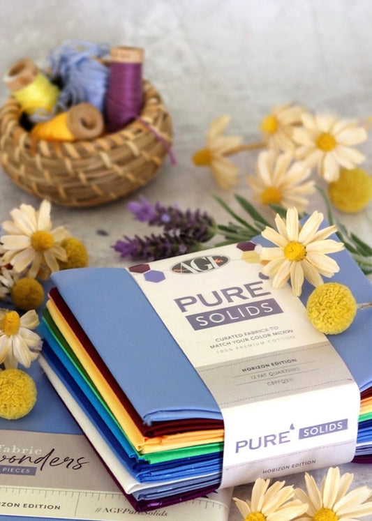 A stack of folded fabric bolts wrapped in a paper band labeled 'PURE SOLIDS' and '12 Color Edition,' styled with daisies, lavender, and a small basket of yarn on a light surface.