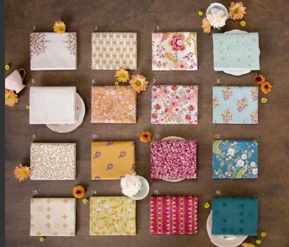 A flat-lay grid of 16 folded fabric fat quarters with various floral, geometric, and solid patterns, arranged on a dark brown wooden background with small decorative flowers and teacups.