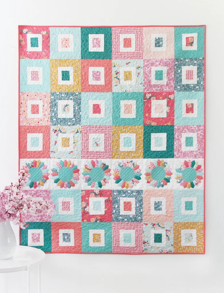 A colorful patchwork quilt hanging on a white wall, featuring a grid of square blocks in pink, teal, yellow, and white floral fabrics, with a floral border and a pink-and-white floral section near the bottom left.