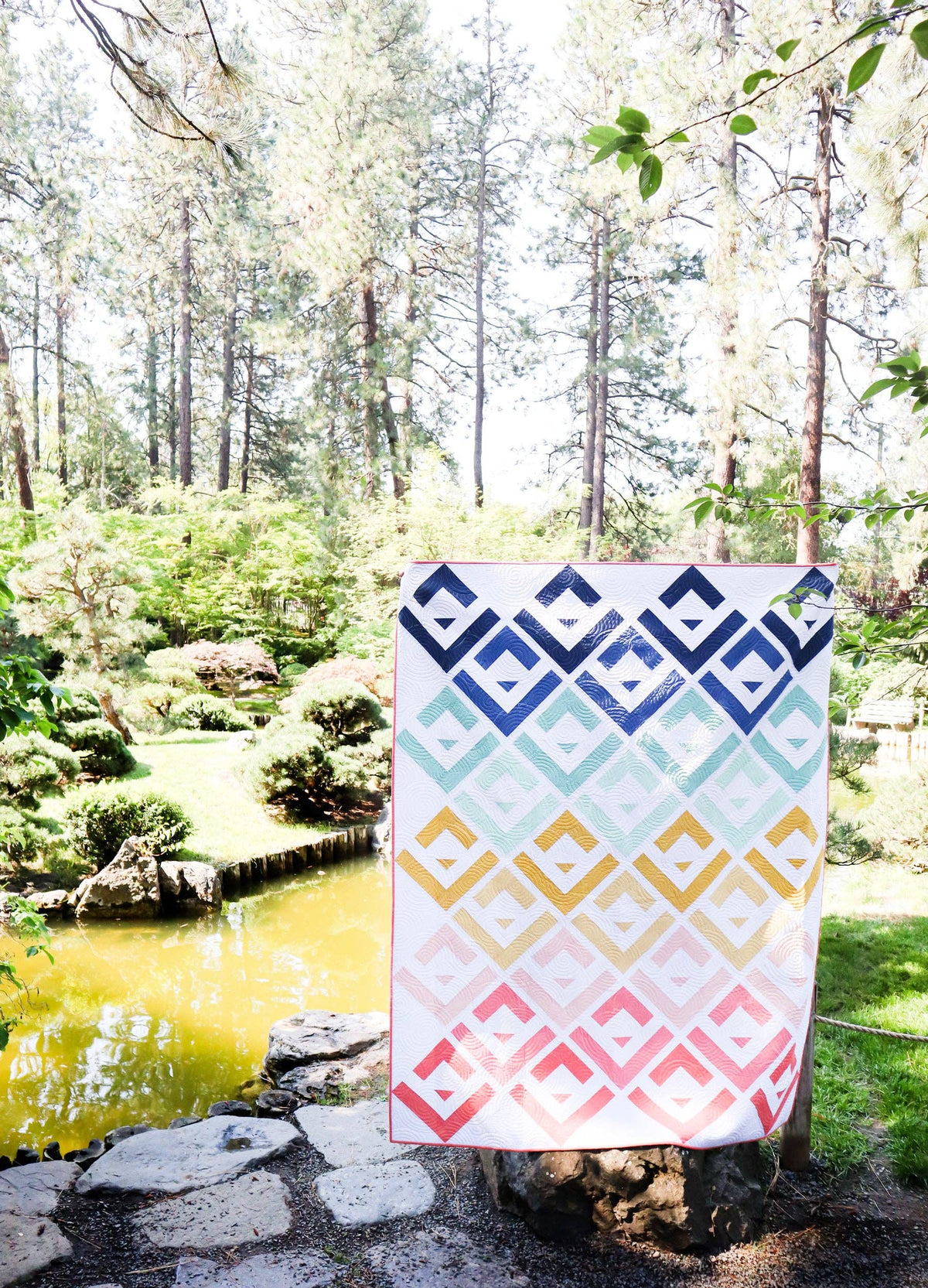 Colorful geometric-patterned quilt draped over a tree stump in a park setting with trees and water in the background.