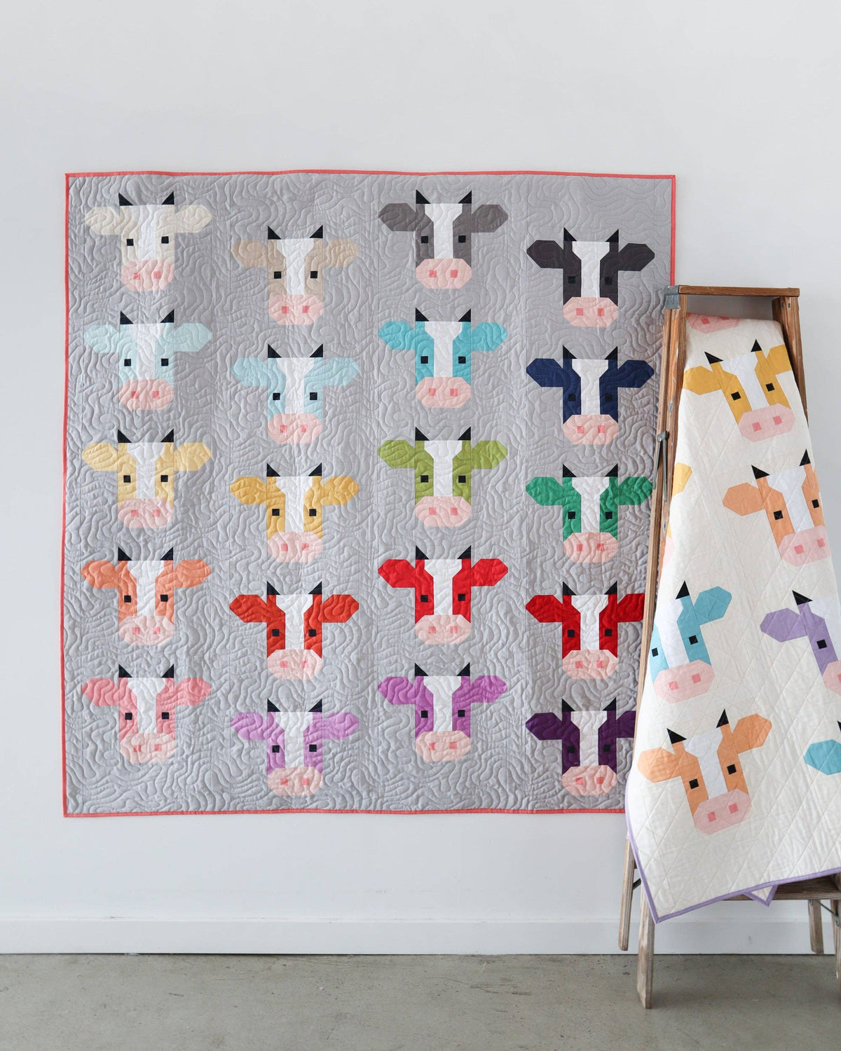 A quilted wall hanging or blanket featuring a grid of colorful cow faces on a light gray background with a pink border, displayed against a white wall with a wooden stand holding a matching folded version.