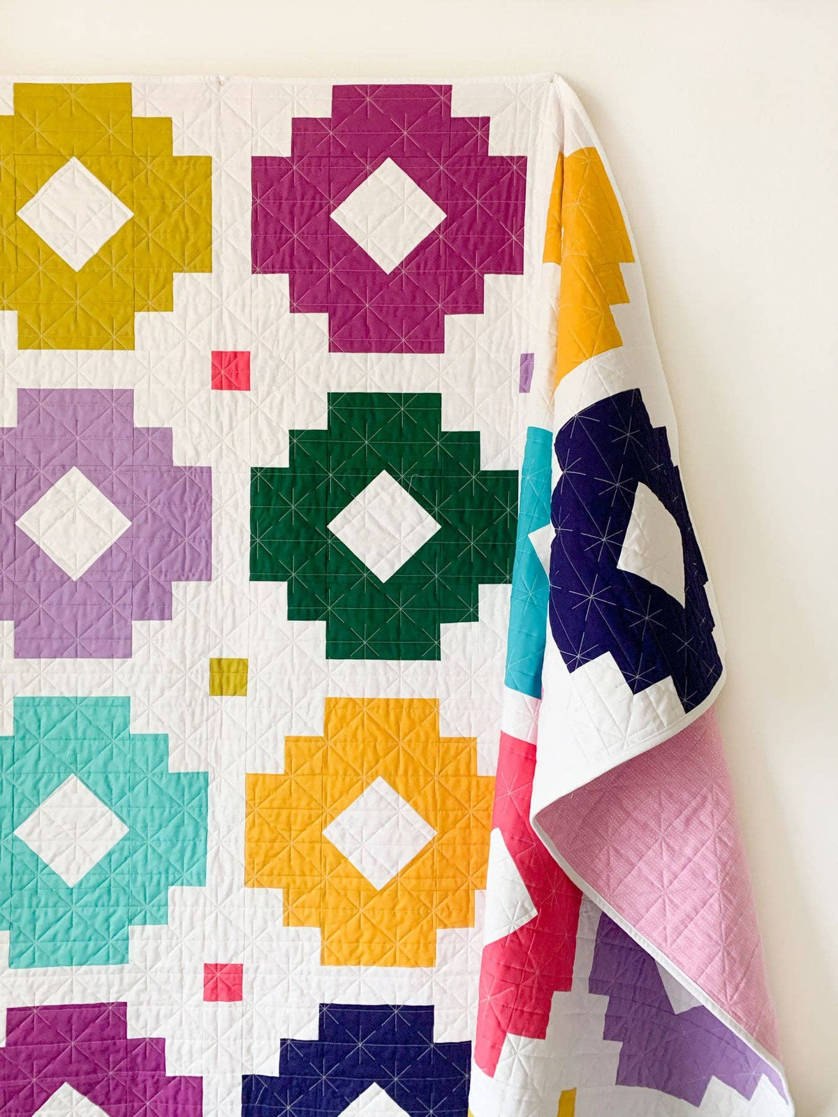 A colorful geometric quilt hanging against a plain light wall, featuring large square motifs in yellow, purple, green, teal, pink, and navy on a white background.