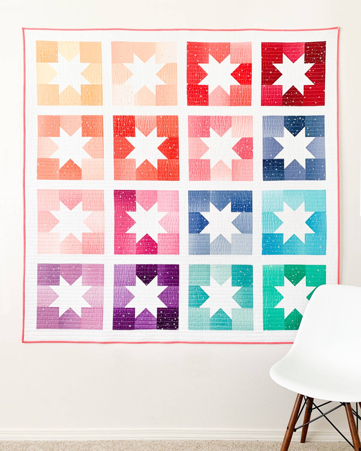 A colorful quilted wall hanging or tapestry with a grid of star-block patterns in peach, coral, red, blue, purple, and teal, displayed on a white wall above a white chair.