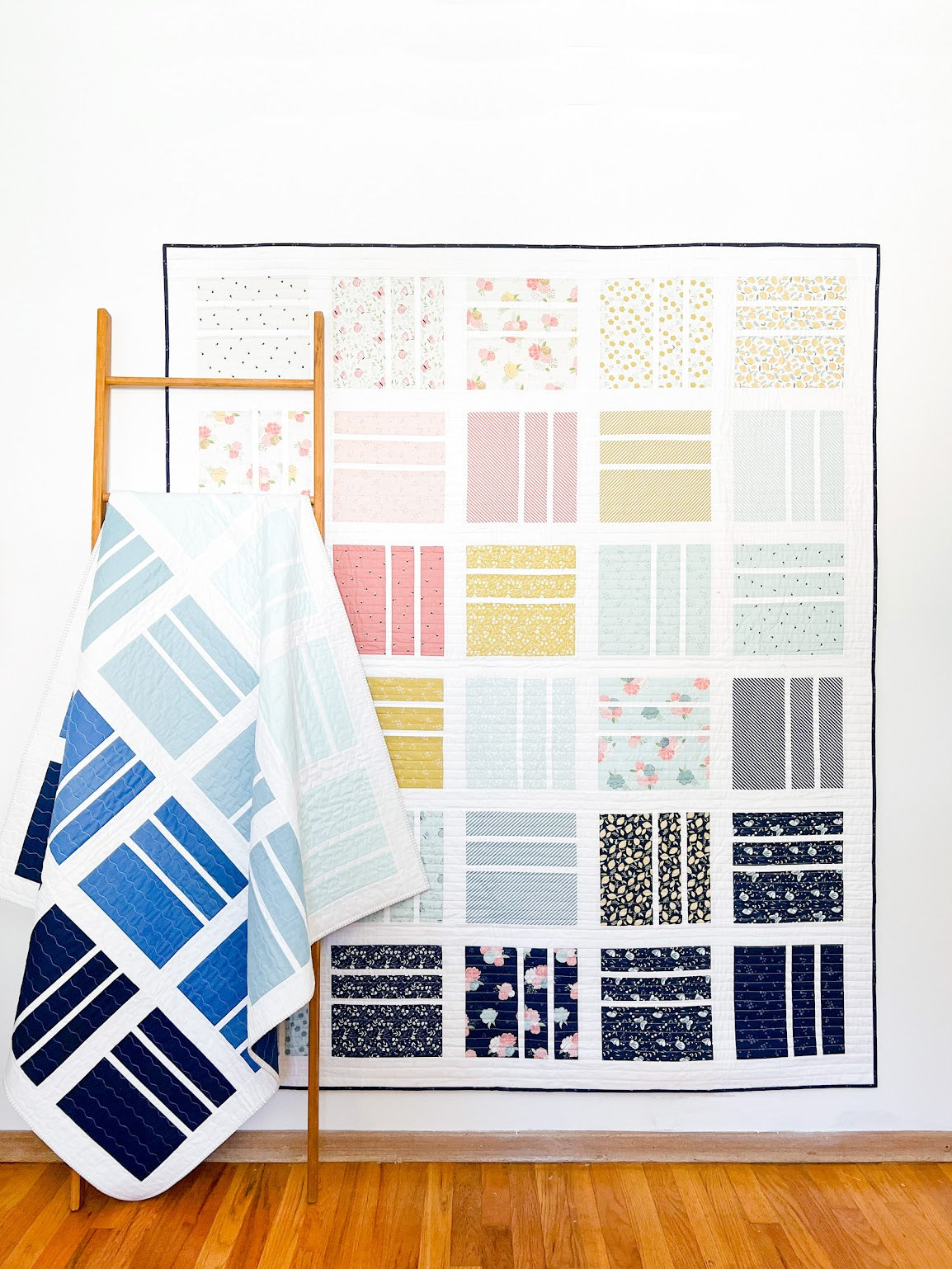 Quilt with various patterns displayed on a wall, with a folded blanket on a wooden ladder.