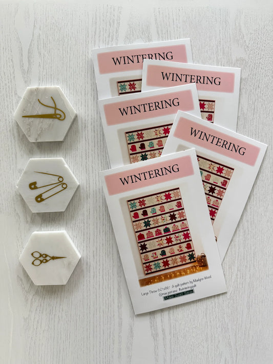 Three printed pattern cards titled WINTERING are arranged on a light wood surface, with three white hexagonal metal craft tools placed to the left. The cards show colorful winter-themed quilt patterns and the brand name 'The Quilted Life' at the bottom.